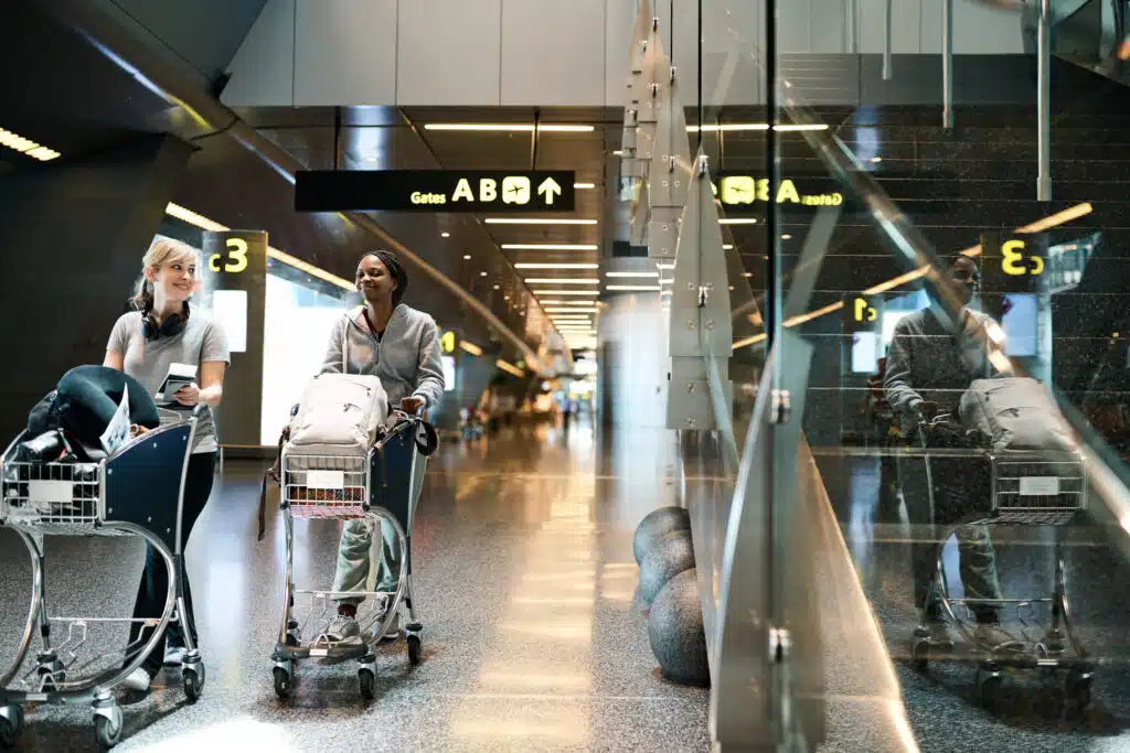Twee jonge vrouwen met bagagekarren in een moderne luchthavenomgeving, klaar om goedkoop te vliegen vanuit België.